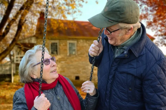 Zwei ältere Personen auf einer Schaukel vor einem Backsteinhaus im Herbst. Sie schauen sich lächelnd an und sind herbstlich mit Mütze und Schal gekleidet.