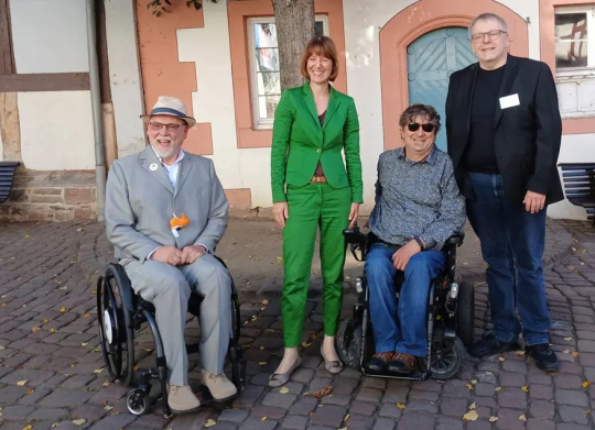 Zwei Männer im Rollstuhl und zwei stehende Personen lächeln auf einem gepflasterten Platz vor einem Fachwerk und Rundbogenfenster in die Kamera. 
