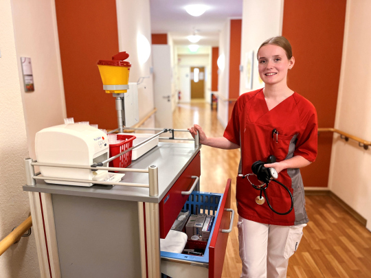 Das Foto zeigt Michelle, eine junge Frau, die eine Ausbildung in der Pflege macht. Sie steht neben einem Medikamentenwagen im Flur des Pflegeheimes und hält ein Blutdruckmessgerät in der Hand. 
