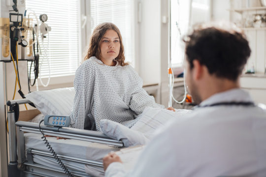 Eine junge Frau sitzt in einem Krankenhausbett. Ein Arzt sitzt vor ihr auf einem Stuhl und hat die Patientenakte in der Hand. 