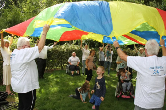 Erwachsene und Kinder heben in einem Park ein großes, buntes Schwungtuch, während Kinder darunter spielen.