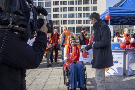 Menschen bei einer Veranstaltung im Freien, eine Person im Rollstuhl trägt eine orangefarbene Warnweste und wird von einem Mann interviewt. Im Hintergrund sind weitere Teilnehmer zu erkennen.