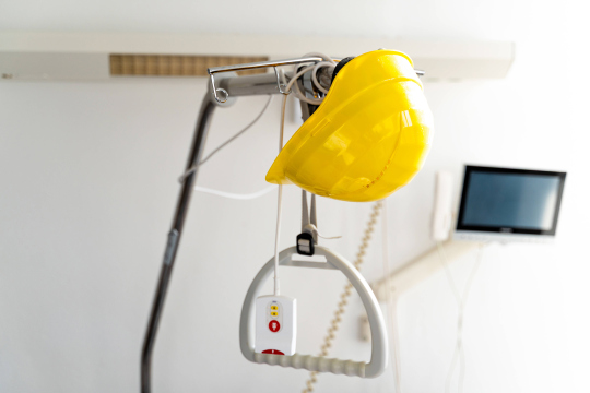 Ein gelber Bauhelm hängt an einem Haltegriff über einem Krankenhausbett; im Hintergrund sind eine Klinikwand und ein Bildschirm zu sehen.