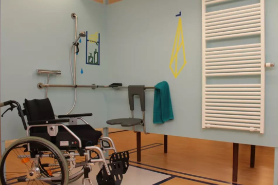 Barrierefreies Badezimmer mit Duschbereich, Haltegriffen und Duschsitz; daneben steht ein Rollstuhl, an der Wand hängt ein Handtuch und ein Heizkörper ist zu sehen.